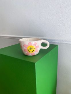 kop med hank og en stor lyserød blomst med gul midte og smiley udenpå. Koppen er håndlavet hos Posy Ceramics.