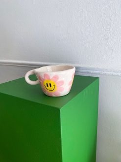 kop med hank og en stor lyserød blomst med gul midte og smiley udenpå. Koppen er håndlavet hos Posy Ceramics.